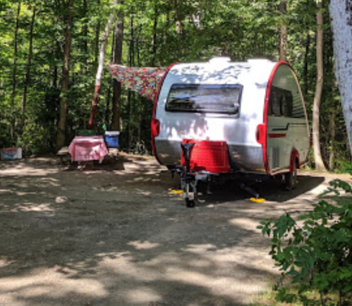A small camper parked in a wooded area, with a picnic table and colorful fabric draped nearby.