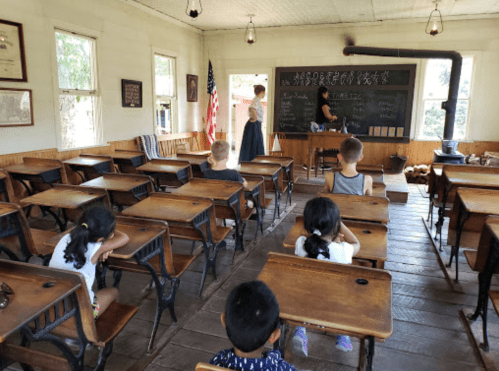 A vintage classroom with students seated at wooden desks, while a teacher writes on a chalkboard. An American flag is visible.