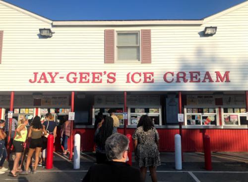 A crowd lines up outside Jay-Gee's Ice Cream shop, featuring a white building with red accents and a menu display.