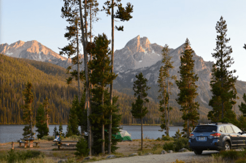 A serene campsite by a lake, surrounded by tall trees and mountains, with a car parked nearby at sunset.