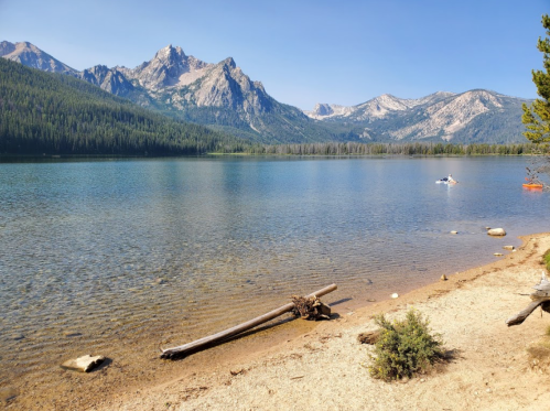 A serene lake surrounded by mountains, with clear water and a sandy shore, featuring kayakers in the distance.