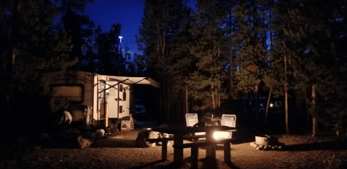 A campsite at night with a camper, two chairs, a fire pit, and trees surrounding the area.