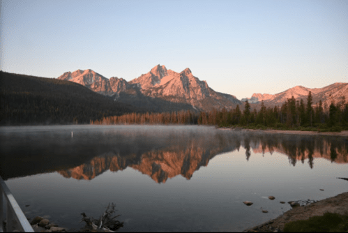 Serene lake at sunrise, reflecting mountains and trees, with mist rising from the water's surface.