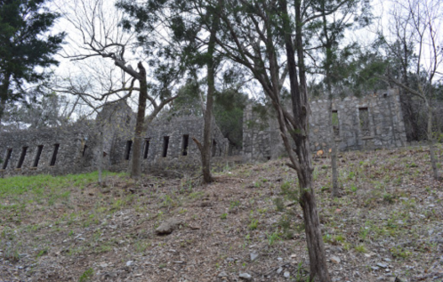 Ruins of a stone building surrounded by trees and rocky terrain, with a grassy area in the foreground.