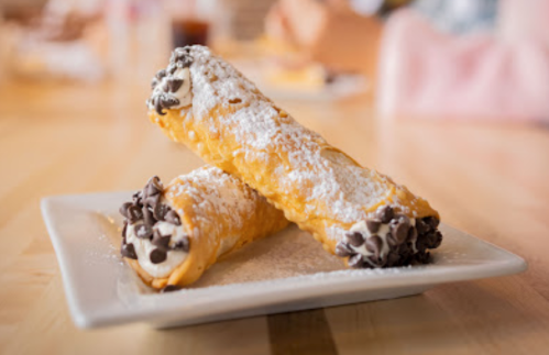 Two chocolate chip cannoli dusted with powdered sugar on a white plate, set on a wooden table.