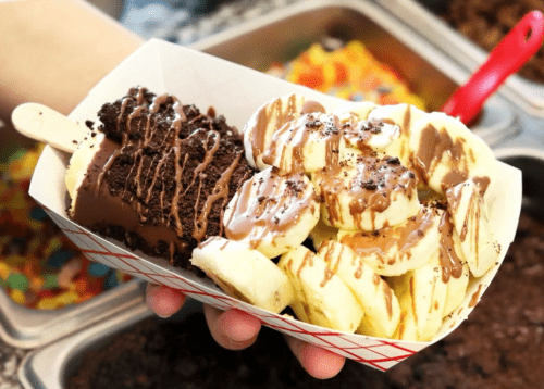 A hand holding a tray with chocolate-covered bananas and a chocolate-coated ice cream bar, drizzled with chocolate sauce.