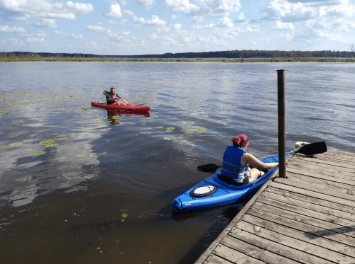 Two people kayaking on a calm lake, one in a red kayak and the other in a blue kayak, with a wooden dock nearby.