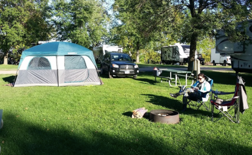 A campsite with a tent, a person sitting in a chair, and a fire pit, surrounded by trees and RVs in the background.