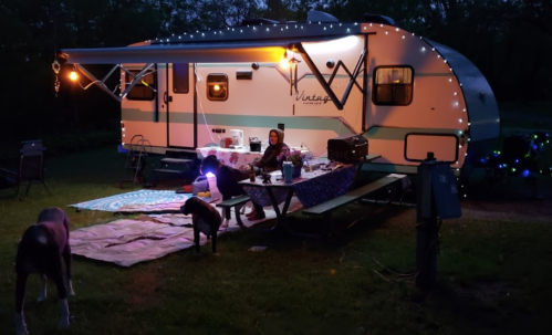 A cozy campsite at night with a vintage trailer, string lights, and a person sitting by a picnic table with dogs nearby.