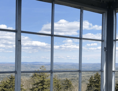 View from a lookout tower, featuring a panoramic landscape of mountains and blue sky with fluffy clouds.
