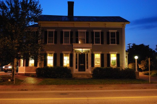 A historic building with dark shutters and illuminated windows, set against a twilight sky.