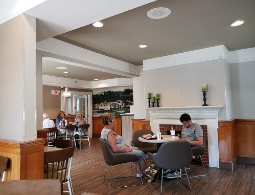 A cozy café interior with patrons seated at tables, a mural on the wall, and a fireplace in the background.
