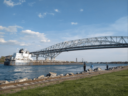 A large ship passes under a steel bridge, with fishermen along the riverbank on a sunny day.