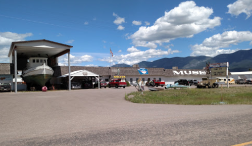 A museum building with a boat on display, surrounded by vintage vehicles and mountains under a blue sky.