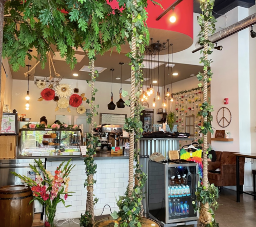 A cozy café interior with greenery, a swing, and a counter displaying baked goods and drinks. Warm lighting enhances the ambiance.