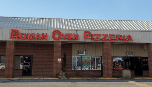 Exterior of Roman Oven Pizzeria, featuring a red sign and a brick facade in a shopping plaza. Clear blue sky above.