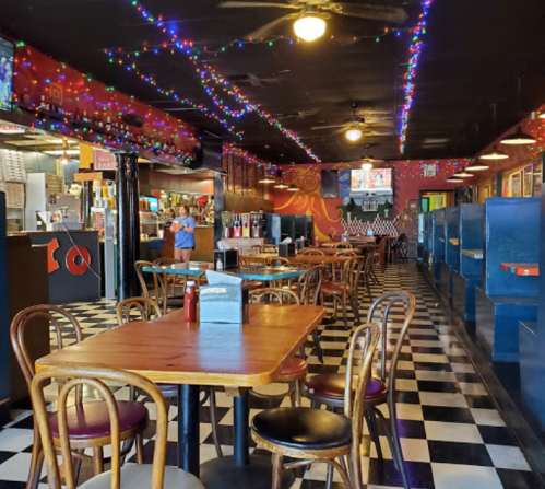 A colorful diner interior with checkered floors, string lights, and empty tables. A staff member is at the counter.