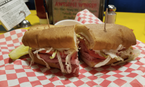 A sandwich with layers of meat, lettuce, and tomato, served on a checkered paper plate with a pickle on the side.