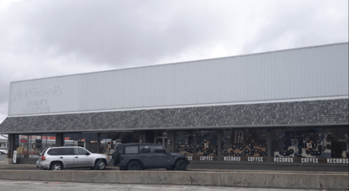A storefront with large windows displaying signs for coffee and records, featuring a gray exterior and parked cars.