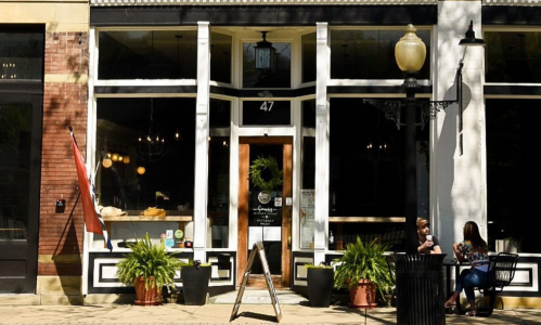 A storefront with large windows, a welcoming entrance, and outdoor seating surrounded by greenery.
