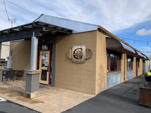 Exterior of a restaurant with a wooden sign, patio seating, and a blue sky in the background.