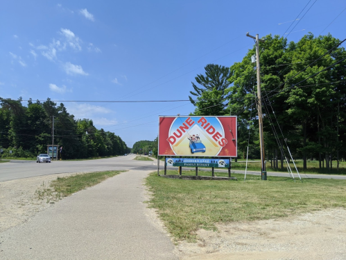 A roadside billboard advertising "Dune Rides" with a blue dune buggy graphic, surrounded by trees and a clear sky.