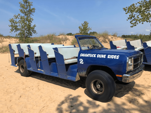 A blue open-air vehicle with bench seating parked on sandy dunes, labeled "Saugatuck Dune Rides."