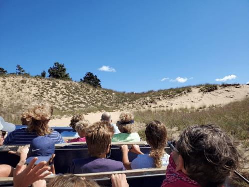 A group of people in a vehicle, facing a sandy dune under a clear blue sky with a few clouds.
