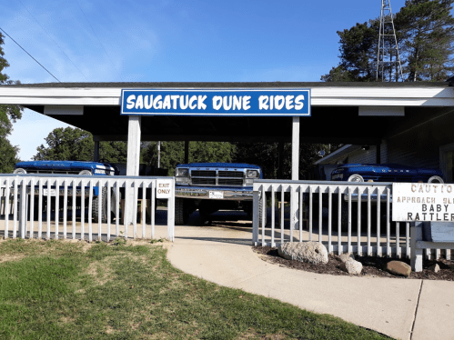 Sign for Saugatuck Dune Rides with blue vehicles parked underneath, surrounded by a white fence and grassy area.