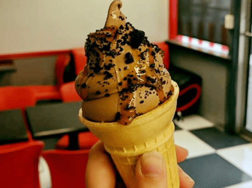 A hand holding a chocolate soft serve ice cream cone topped with crushed cookies and drizzled with caramel.