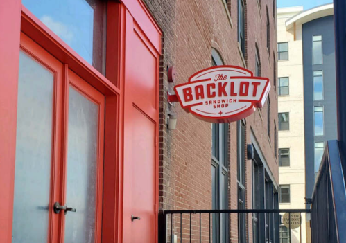 Sign for "The Backlot Sandwich Shop" on a brick building with red doors.