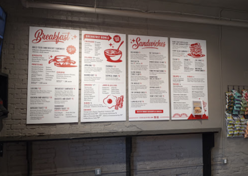 Menu board featuring breakfast and sandwich options, with prices and descriptions, displayed on a brick wall.