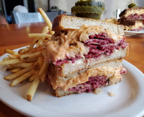 A stacked sandwich with layers of pastrami and melted cheese, served with fries and topped with a pickle.