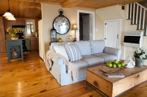 Cozy living room with a light blue couch, wooden coffee table, and a kitchen in the background. Sunlight fills the space.