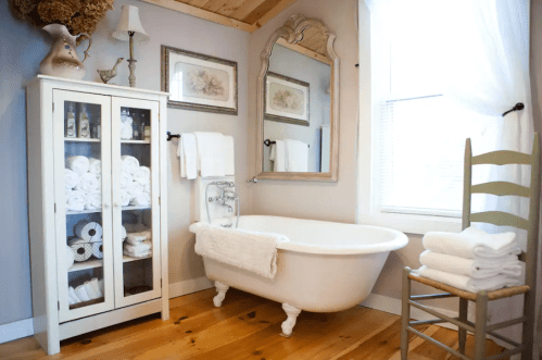 A cozy bathroom featuring a freestanding tub, a glass cabinet with towels, and a wooden chair. Natural light filters in.