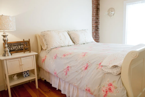 Cozy bedroom with a floral bedspread, bedside table, lamp, and exposed brick wall. Natural light from a window.