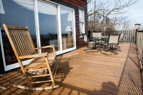 A wooden rocking chair and a dining set on a sunlit deck with a view of trees and a distant landscape.