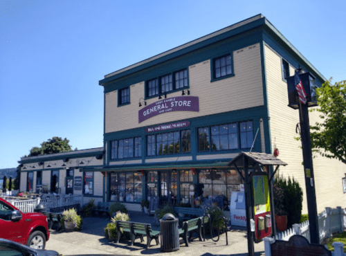A charming general store with a green and yellow exterior, featuring a sign and outdoor seating. Clear blue sky above.