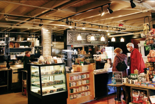 A cozy café interior with a display case, menu board, and customers browsing. Warm lighting and rustic decor.
