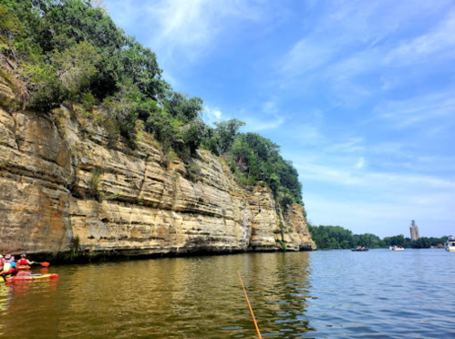 A serene river scene with rocky cliffs and lush greenery under a blue sky, with kayakers in the foreground.