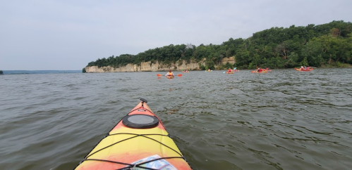 A kayak in the foreground with a view of a calm river and several kayakers in the distance near a wooded shoreline.