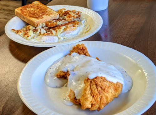 A plate of fried chicken with gravy, alongside a plate of eggs, hash browns, and toast on a wooden table.