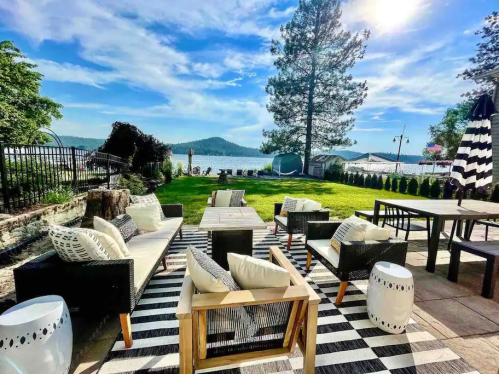 A scenic outdoor seating area with black and white furniture, overlooking a lake and mountains under a bright blue sky.
