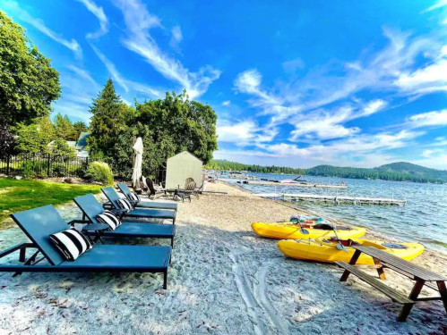 Lounge chairs on a sandy beach by a lake, with kayaks and a clear blue sky in the background.