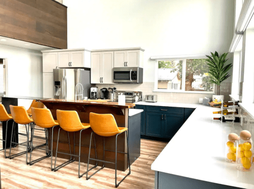Modern kitchen with a large island, orange bar stools, stainless steel appliances, and bright natural light.