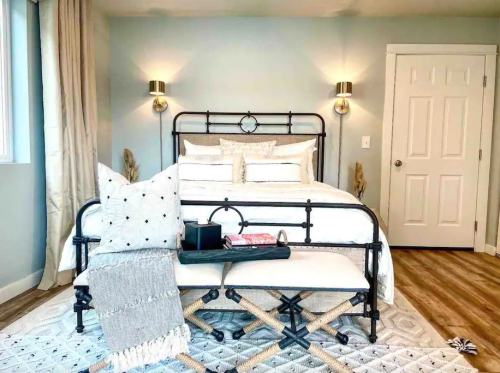 A cozy bedroom featuring a metal bed frame, decorative pillows, a bench, and soft lighting against a light blue wall.