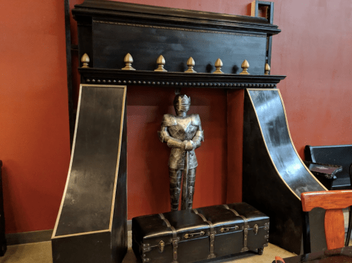 A decorative fireplace with a knight statue and a wooden chest beneath it, set against a red wall.