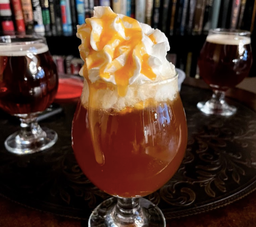 A glass of caramel-topped dessert drink with whipped cream, set against a backdrop of books and other drinks.