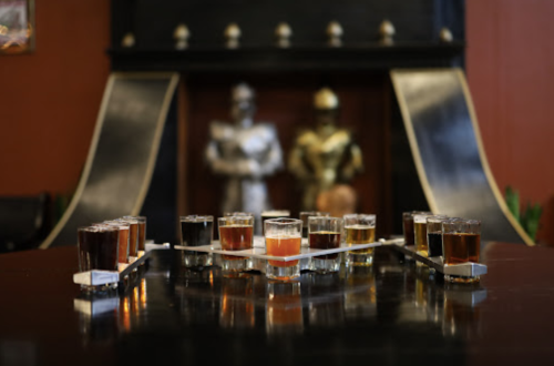 A row of shot glasses filled with various colored liquids on a table, with two knight statues in the background.