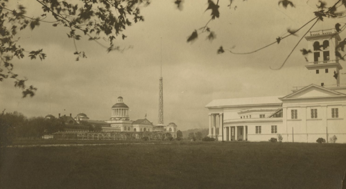 Historic black-and-white photo of a grand building complex with a tall tower, surrounded by trees and open grass.